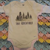 Baby onesie with 'Tiny Adventurer' text and tree design on a colorful crocheted blanket