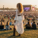 A woman with long curly hair stands on a grassy field at an outdoor concert, seen from behind, wearing a white bohemian lace maxi dress with a brown belt and brown boots, with a large crowd and stage in the background during golden hour.